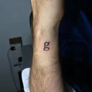 Фото минималистичной татуировки с буквой 'g' на запястье. Простой и лаконичный дизайн, выполненный черными чернилами. Подходит для тех, кто ценит сдержанность и индивидуальность.
