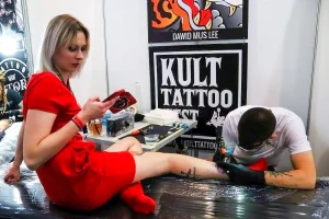 Фото тату процесса нанесения татуировки девушке на ногу. Мастер работает, а клиент сидит в кресле и смотрит в телефон.