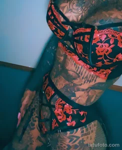 Фотография девушки с татуировками на теле, одетой в прозрачный топ с цветочным узором. Татуировки выполнены в темных тонах и имеют разнообразный характер.