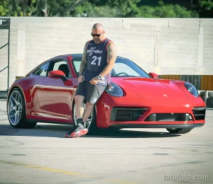 Фотография мужчины с татуировками, стоящего рядом с красным Porsche. Мужчина одет в спортивную одежду и смотрит в камеру.