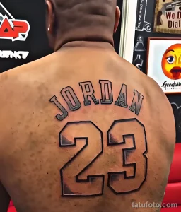 Фотография крупным планом татуировки с надписью "JORDAN" и номером "23" на спине мужчины. Детализированный рисунок, спортивная тематика.