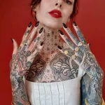 Фотография девушки с множеством татуировок на шее, руках и теле. Она одета в серую футболку и украшения.