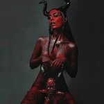 Фотография девушки с яркой красной татуировкой, изображающей демоническую тематику. На голове рога, а на теле - сложные узоры и рисунки.