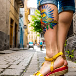 Как вес влияет на тату A petite woman with a colorful bird tattoo on her ankle walking along a cobblestone street wearing summer sandals medium-leng tatufoto.com 169