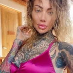 Фотография девушки с татуировками на лице, шее, груди и руках. Татуировки выполнены в ярких цветах и графичном стиле. Девушка позирует в розовом топе.
