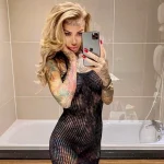 Фотография девушки с обилием татуировок на теле, в черном платье, делающей селфи в ванной комнате. Тату покрывают руки, ноги и шею.
