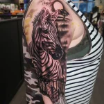 Фото тату: красочная татуировка головы зебры на плече, с изображением африканского пейзажа и силуэтами деревьев на фоне заката.