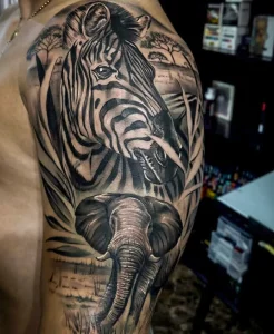 Фото тату: реалистичное изображение головы зебры и слона на плече, выполненное в черно-серых тонах с элементами пейзажа.