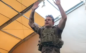Фото мужчины с татуировкой 'Deus Vult' и крестом на руке. Мужчина в тактической экипировке, с поднятыми руками.