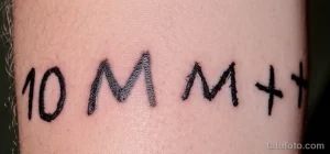 Фото тату с минималистичным дизайном: цифры '10', буквы 'M', 'M' и символ '+'. Черный рисунок на руке, простой и лаконичный.
