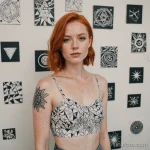 pale redhead woman with freckles and subtle vitiligo on collarbone, standing next to wall covered with tattoo flash sheets, wear 2025 №46 - фото tatufoto.com