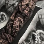 Фото тату октопуса, обвивающего руку, с детально прорисованным старинным кораблем на фоне. Черно-белая татуировка в реалистичном стиле.
