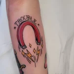 Фото тату магнита с надписью "Trouble" на руке. Рисунок выполнен в стиле олдскул, с четкими контурами и яркими цветами.