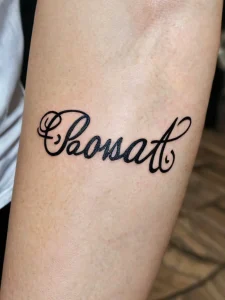 Фото татуировки на предплечье с надписью 'Rosat', выполненной в красивом каллиграфическом стиле. Черно-белый рисунок татуировки.