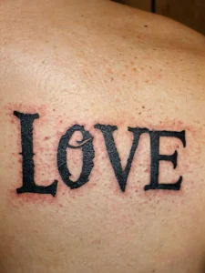 Фото тату: надпись 'LOVE' черным цветом на лопатке. Простой и лаконичный дизайн, классический шрифт.