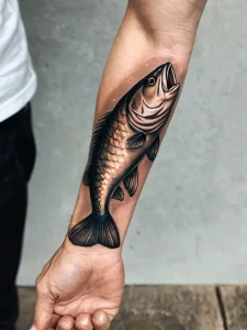Фото тату с изображением рыбы, выполненной в классическом стиле на предплечье. Черно-белый рисунок татуировки с реалистичными деталями и четкими контурами.