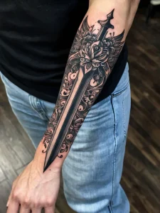Фото татуировки с изображением меча, окруженного цветами. Черно-белый рисунок, выполненный в детализированном стиле с использованием графических элементов.
