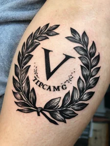 Фото татуировки с изображением лаврового венка и инициалов 'V' и 'TIPCAMG'. Черно-белый рисунок, выполненный в стиле графика, создает стильный и индивидуальный образ.