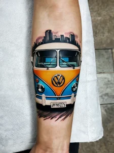 Фото тату: яркий рисунок Volkswagen Bus на предплечье с изображением городского пейзажа на фоне.