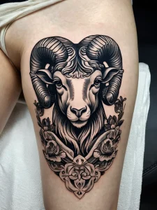Фото татуировки головы барана, окруженной цветами, на бедре. Черно-белый рисунок с детализированной проработкой шерсти, рогов и лепестков. Элегантный и женственный дизайн.