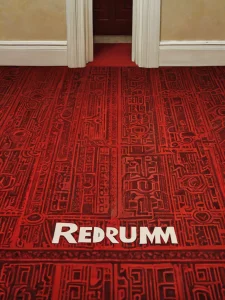 Фото красного ковра с геометрическим узором и надписью 'Redrum', отсылка к фильму 'Сияние'. Мрачная и атмосферная композиция.