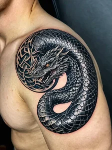 Черно-серая татуировка на плече с изображением дракона, свернувшегося в спираль, окруженного кельтским узором, выполненная в реалистичном стиле.