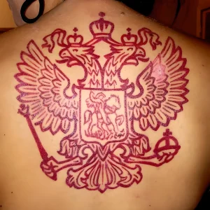 Фото татуировки на плече, изображающей двуглавого орла в красных тонах. Рисунок выполнен в технике контура, с использованием градиентов и теней.