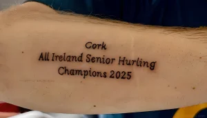 На руке изображена татуировка с надписью 'Cork All Ireland Senior Hurling Champions 2025' (Корк - Чемпионы Высшей лиги по хёрлингу Ирландии 2025).
