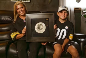 Женщина и мальчик, одетые в форму Pittsburgh Steelers, держат в руках рамку с изъятым и законсервированным фрагментом татуировки