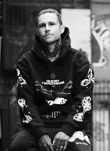 Corey Divine, тату-мастер, портрет в черном худи с геометрическими татуировками на руках и шее.