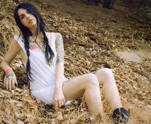 Пэнди, модель SuicideGirls, с татуировками на руках и ногах, сидящая на фоне осеннего пейзажа.