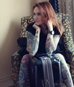 Леа Вендетта, тату-модель, с татуировками на руках и ногах, сидящая с чемоданом.