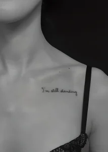 Черно-белая фотография татуировки надписи 'I'm still standing' на ключице девушки. Минималистичный стиль, тонкие линии.