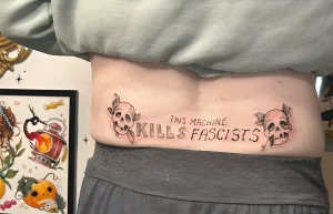 Татуировка на пояснице с черепами и надписью 'This machine kills fascists'