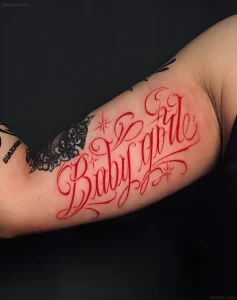 Крупный план татуировки надписи "Baby Girl" красным цветом на плече девушки, с изображением крыльев бабочки.