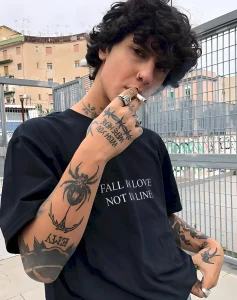 Молодой человек с кудрявыми волосами, в черной футболке с надписью "FALL IN LOVE NOT IN LINE", курит сигарету и демонстрирует татуировки на руках, шее и груди, включая изображение паука.