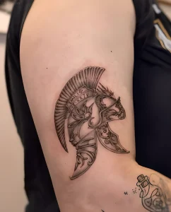 Черно-белая татуировка на плече с изображением шлема спартанца, украшенного растительным орнаментом и римскими цифрами.