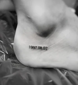 Черно-белая татуировка даты '1997.08.02' на внутренней стороне стопы, выполненная четким шрифтом.