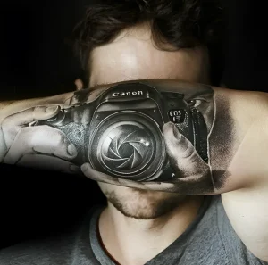 Черно-белая реалистичная татуировка фотоаппарата Canon EOS на предплечье, создающая иллюзию, будто камера является продолжением руки.