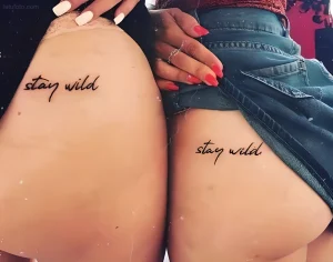 Два женских бедра, на которых нанесены идентичные татуировки надписью "stay wild", выполненной изящным рукописным шрифтом. Татуировки расположены сбоку от талии.
