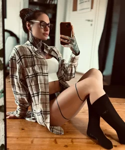 Модель joii_xoxo в клетчатой рубашке и черных гольфах, делающая селфи в зеркале, демонстрируя татуировки на руках и шее.