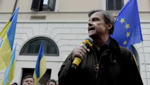Итальянский сенатор Карло Календа выступает с микрофоном на фоне украинского и европейского флагов.