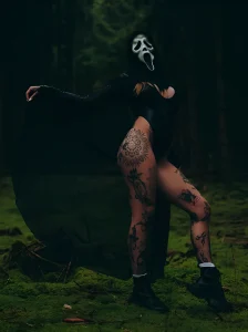 Aline mc.lovin.ink в маске и черном плаще, демонстрирующая татуировки на ногах в лесу