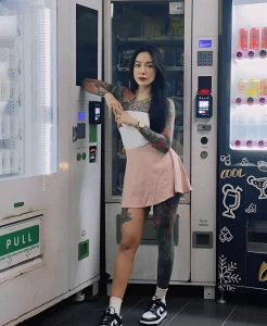 Janejaneey у торгового автомата, демонстрирующая свои татуировки на руках и ногах.