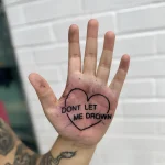Черно-белая татуировка на ладони с надписью 'DON'T LET ME DROWN' внутри контура сердца, выполненная в минималистичном стиле.