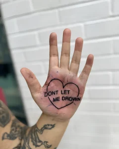 Черно-белая татуировка на ладони с надписью 'DON'T LET ME DROWN' внутри контура сердца, выполненная в минималистичном стиле.