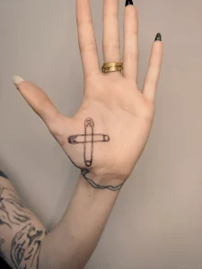 Черно-белая татуировка булавки на ладони, выполненная в технике тонкой линии, на женской руке с золотым кольцом.