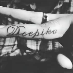 Черно-белое фото татуировки на пальце с именем 'Deepika', выполненной каллиграфическим шрифтом.