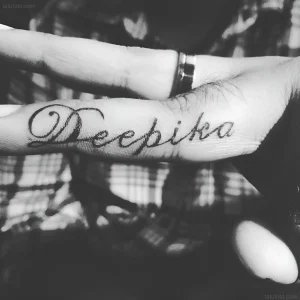 Черно-белое фото татуировки на пальце с именем 'Deepika', выполненной каллиграфическим шрифтом.