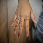 Фотография женской руки с минималистичными татуировками на пальцах: маленькие звезды, полумесяц, компас и геометрические линии.